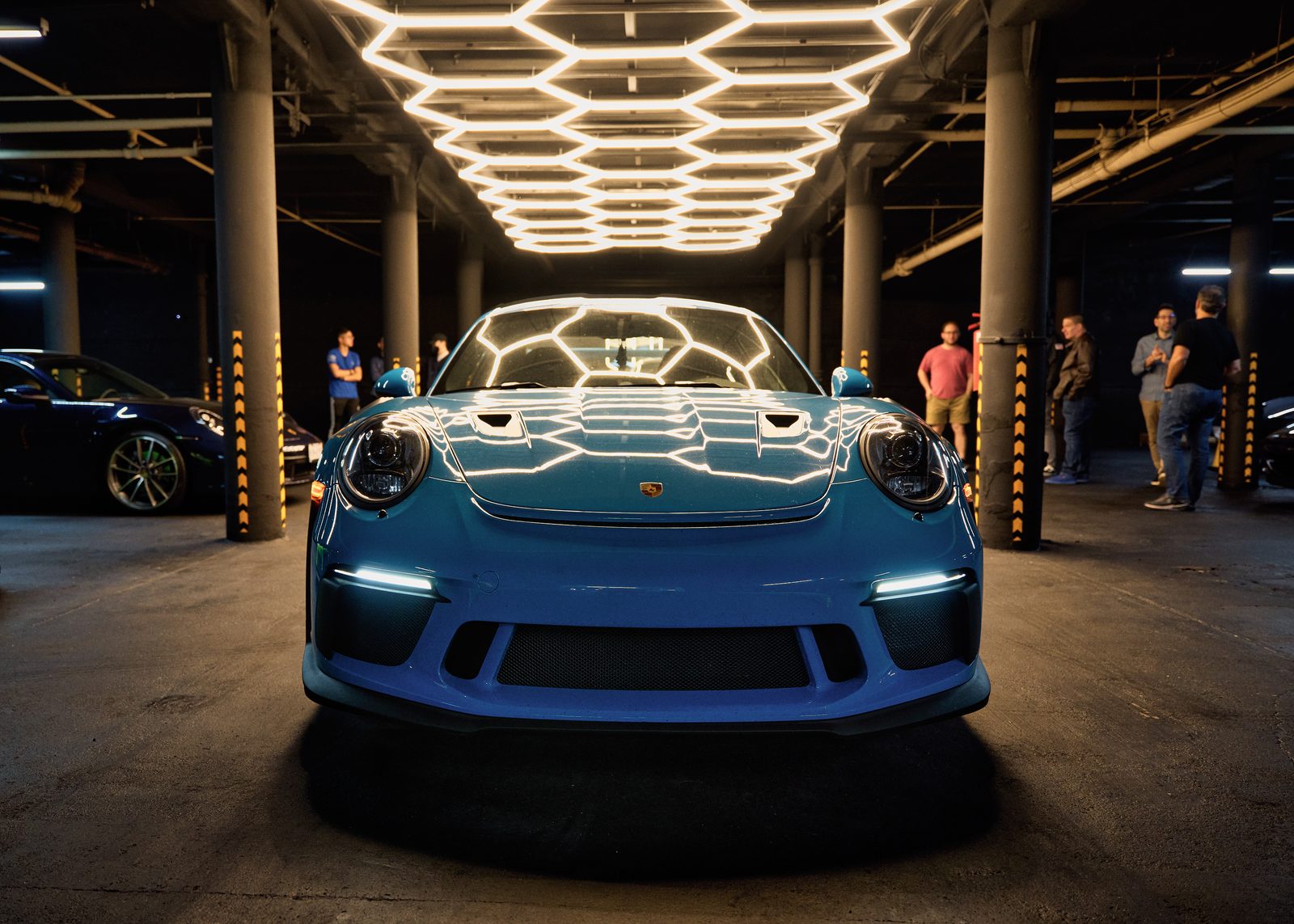 A blue Porsche GT3 head-on under the hexagon light ceiling of the Paddock garage