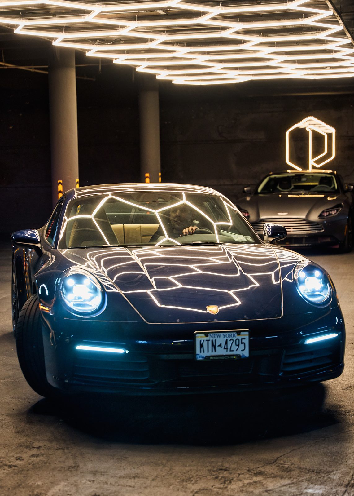 A dark Porsche 911 with the hexagon lights reflected across its bonnet, the Paddock mark lit behind