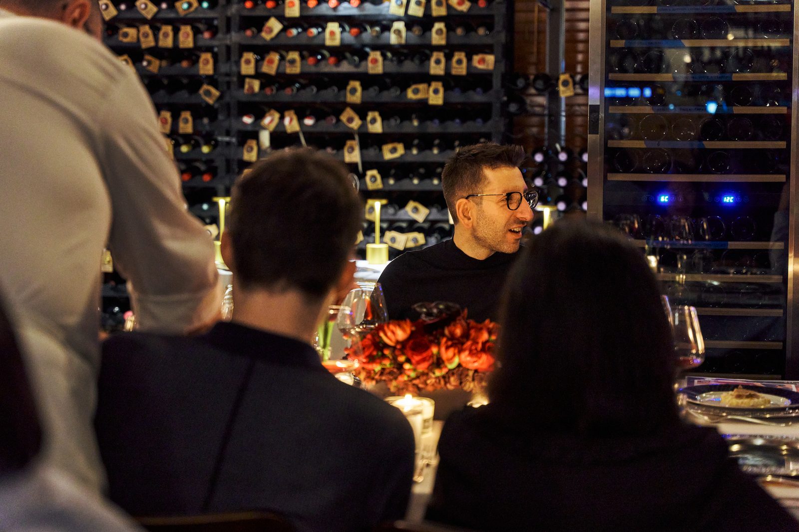 Guests at the table in the wine cellar, seen from behind, red flowers and the wall of bottles beyond