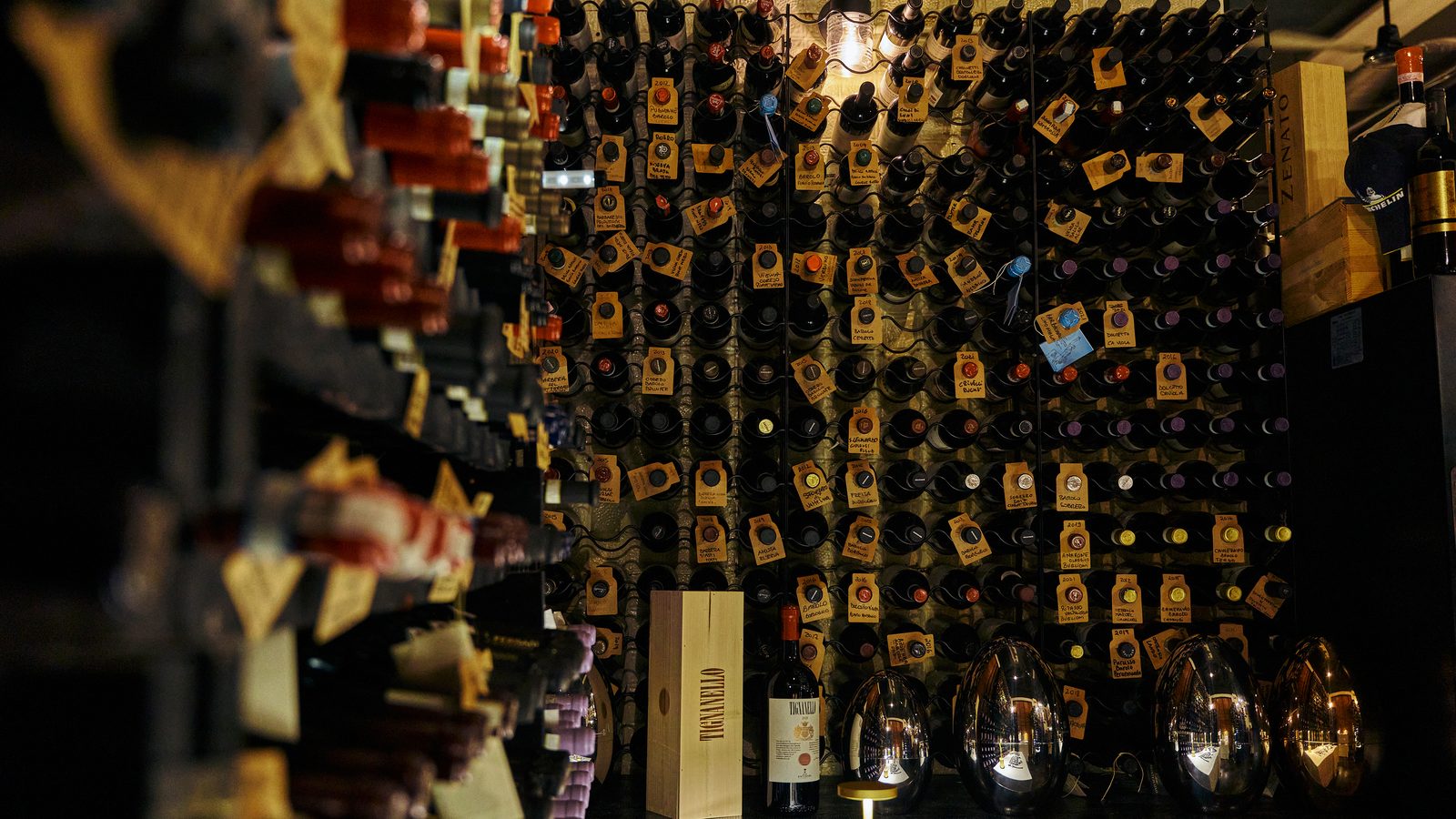 The wall of the wine cellar, bottles racked with handwritten tags, lamps below